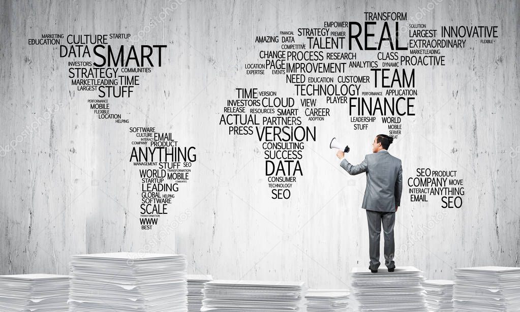 Businessman in suit standing on pile of documents with speaker in hand with business-related terms in form of world map on background. Mixed media.