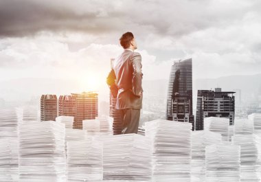 Takım elbiseli kendine güvenen genç işadamı şehir manzarası ve arka planda güneş ışığı olan bir yığın belgenin üzerinde duruyor. Karışık ortam.