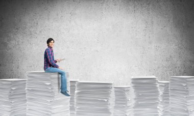 Casual giyim gri arka plan duvara ile belgeler yığını üzerinde oturan genç adam. Karışık teknik.