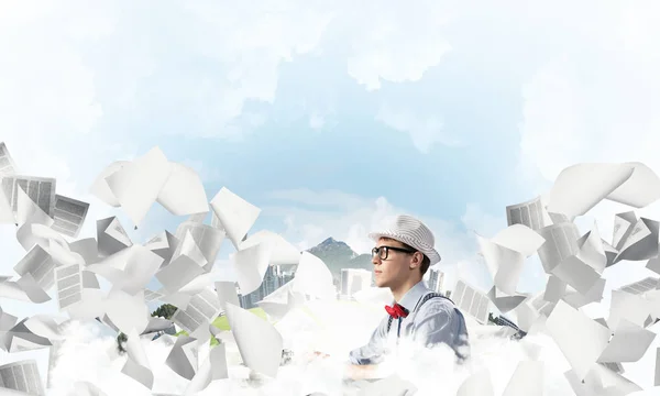 Young man writer in hat and eyeglasses using typing machine while sitting at the table among flying papers with floating city and cloudy skyscape on background.