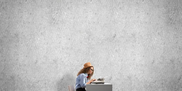 帽子と眼鏡の背景に灰色のコンクリートの壁にテーブルに座ってタイピング マシンを使用しての若い美しい女性作家の側面図 — ストック写真