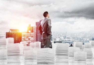 Takım elbiseli kendine güvenen genç işadamı şehir manzarası ve arka planda güneş ışığı olan bir yığın belgenin üzerinde duruyor. Karışık ortam.