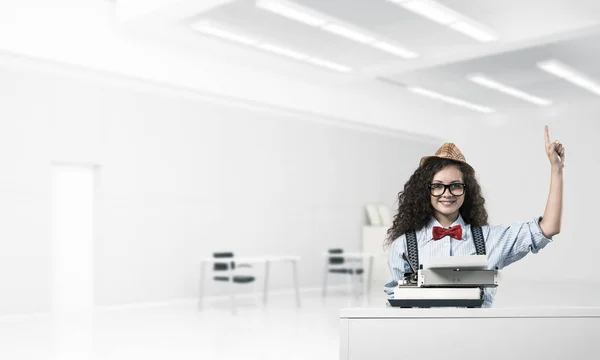 戴着帽子和眼镜的年轻女作家 使用打字机 在坐在桌子上的时候 在办公室的背景下 指向正面 — 图库照片