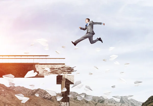 Businessman jumping over gap in bridge among flying paper planes as symbol of overcoming challenges. Skyscape with sunlight and nature view on background. 3D rendering.
