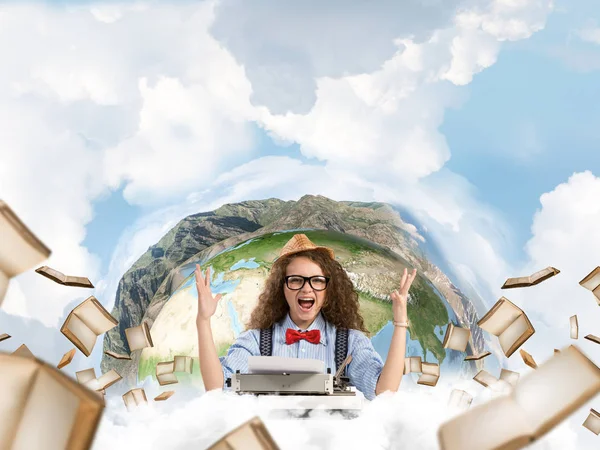 Happy and young woman writer in hat and eyeglasses looking happy while sitting with typing machine at the table with flying books and Earth globe among cloudy skyscape on background. Elements of this image furnished by NASA