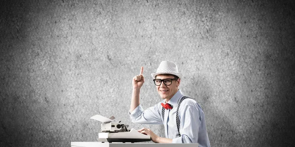Young man writer in hat and eyeglasses using typing machine and pointing upside while sitting at the table against gray concrete wall on background.