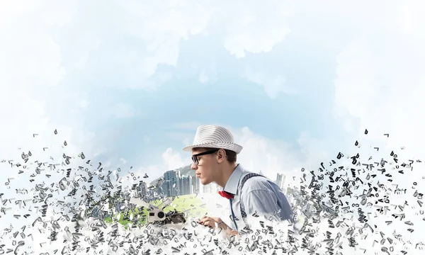 Young man writer in hat and eyeglasses using typing machine while sitting at the table among flying letters with floating city and cloudy skyscape on background.