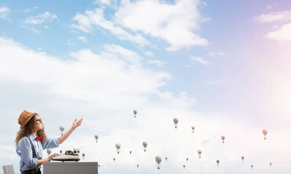 Young and beautiful woman writer in hat and eyeglasses using typing machine while sitting at the table with flying aerostats and cloudy skyscape on background.