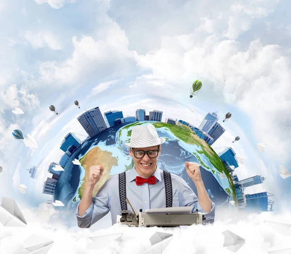 Cheerful young man writer in hat and eyeglasses looking happy while sitting with typing machine at the table with flying paper planes and Earth globe among cloudy skyscape on background. Elements of this image furnished by NASA