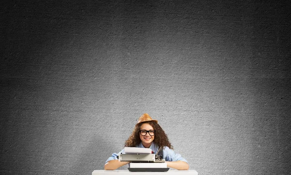 Young and beautiful woman writer in hat and eyeglasses using typing machine while sitting at the table against gray concrete wall on background.