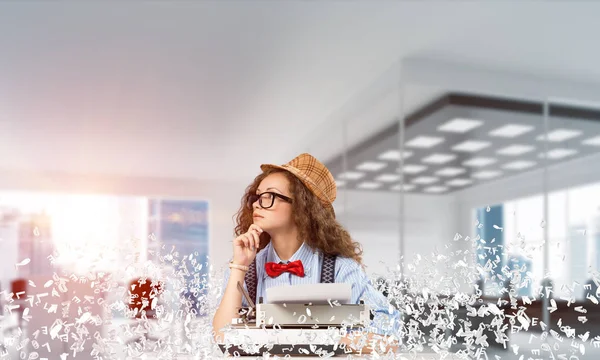Young woman writer in hat and eyeglasses using typing machine while sitting at the table indoors among flying letters and with office view on background.