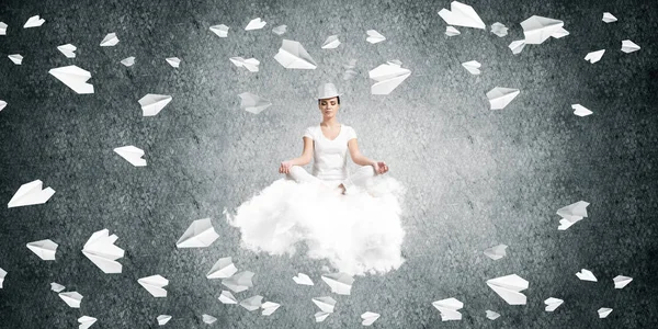 Woman in white clothing keeping eyes closed and looking concentrated while meditating on flying cloud among flying paper planes with gray dark wall on background.