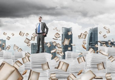 Takım elbiseli kendine güvenen işadamı arka planda şehir manzarası olan uçan kitaplar arasında doküman yığınının üzerinde duruyor. Karışık ortam.