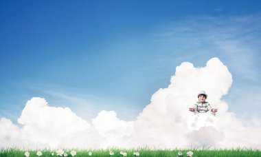 Küçük çocuk gözlerini kapalı tutuyor ve odaklanmış görünüyor. Aynı zamanda bulutlar üzerinde meditasyon yapıyor. Arka planda parlak ve güzel bir manzara var..