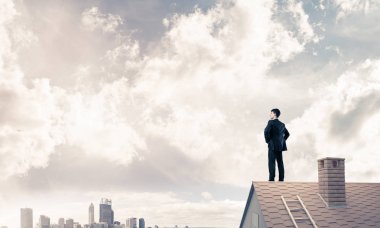 Kararlı genç işadamı evin çatısında dikiliyor ve başka tarafa bakıyor. Karışık ortam