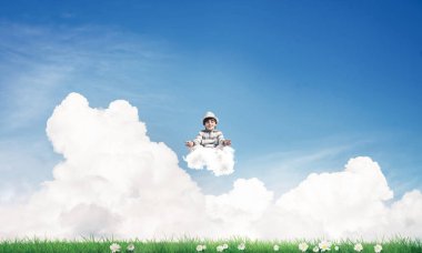 Küçük çocuk gözlerini kapalı tutuyor ve odaklanmış görünüyor. Aynı zamanda bulutlar üzerinde meditasyon yapıyor. Arka planda parlak ve güzel bir manzara var..