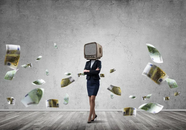 Business woman in suit with old TV instead of head keeping arms crossed while standing among flying euro banknotes inside empty room with gray dark wall on background.