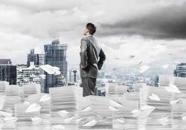 Takım elbiseli kendine güvenen işadamı arka planda şehir manzarası olan uçan kağıt uçaklar arasında evrak yığınının üzerinde duruyor. Karışık ortam.