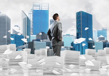 Takım elbiseli kendine güvenen işadamı arka planda şehir manzarası olan uçan kağıt uçaklar arasında evrak yığınının üzerinde duruyor. Karışık ortam.