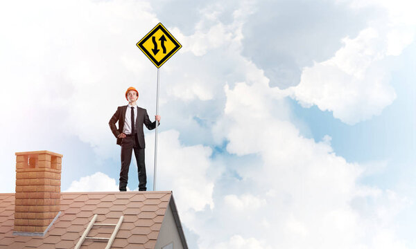 Young determined businessman standing on house roof and showing yellow roadsign. Mixed media
