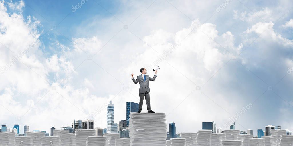 Businessman in suit standing on pile of documents with speaker in hand with skyscape and city view on background. Mixed media.