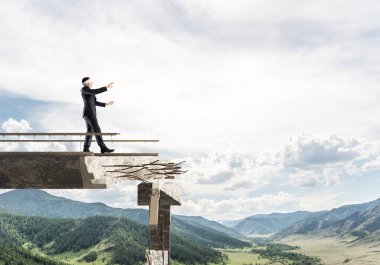 Gizli tehditlerin ve risklerin sembolü olarak beton köprüde gözü kapalı yürüyen bir iş adamı. Arka planda gökyüzü ve doğa manzarası. 3B görüntüleme.