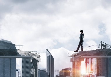 Gizli tehditlerin ve risklerin sembolü olarak beton köprüde gözü kapalı yürüyen bir iş adamı. Arka planda güneş ışığı olan şehir manzarası. 3B görüntüleme.