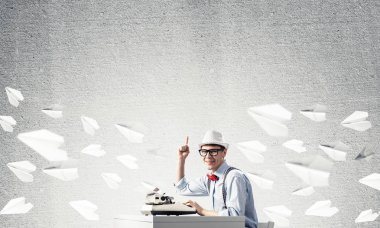 Genç yazar şapka ve gözlük arka plan üzerinde uçan kağıt uçaklar arasında ve gri beton duvara karşı masada otururken yazarak makine ve işaret ters kullanarak adam.