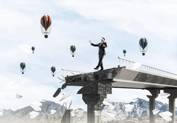 Businessman walking blindfolded among flying paper planes on concrete bridge with huge gap as symbol of hidden threats and risks. Flying balloons and nature view on background. 3D rendering.