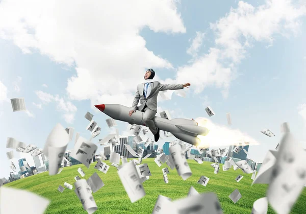 Conceptual image of young businessman in suit flying on rocket among flying papers with cityscape and blue sky on background.