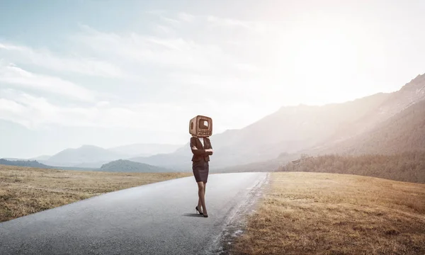 Business woman in suit with an old TV instead of head keeping arms crossed while standing on the road with beautiful landscape on background.