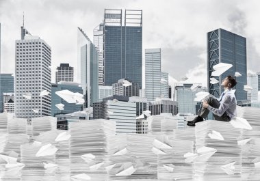 Düşünceli işadamı, arka planda şehir manzarası olan uçan kağıt uçaklar arasında bir yığın belgenin üzerinde otururken başka tarafa bakıyor. Karışık ortam.