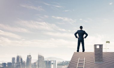 Kararlı genç işadamı evin çatısında dikiliyor ve başka tarafa bakıyor. Karışık ortam