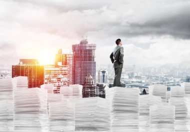 Takım elbiseli kendine güvenen genç işadamı şehir manzarası ve arka planda güneş ışığı olan bir yığın belgenin üzerinde duruyor. Karışık ortam.