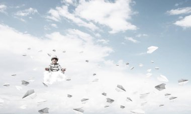 Küçük çocuk gözlerini kapalı tutuyor ve odaklanmış görünüyor. Arka planda bulutlu bir gökyüzü ile havada uçan kağıt uçaklar arasında meditasyon yapıyor..