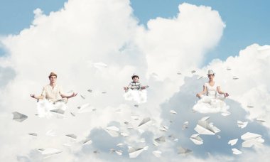 Genç aile gözlerini kapalı tutuyor ve odaklanmış görünüyor. Arka planda bulutlu bir gökyüzü ile havada uçan kağıt uçaklar arasında meditasyon yaparken..