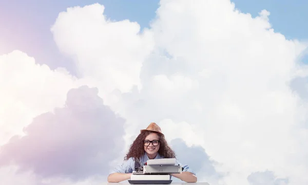 年轻和美丽的女作家在帽子和眼镜使用打字机 而坐在桌子上多云影像在后台 — 图库照片
