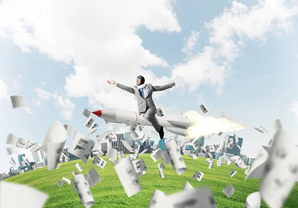 Conceptual image of young businessman in suit flying on rocket among flying papers with cityscape and blue sky on background.