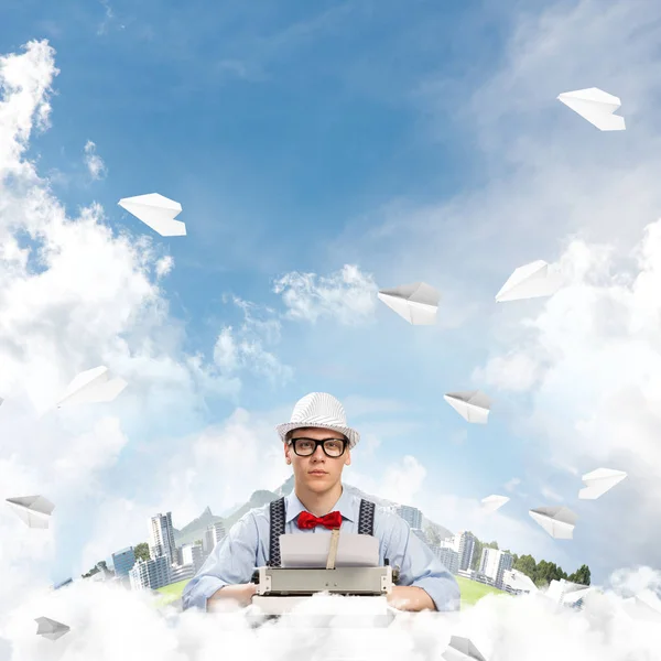 青年作家在帽子和眼镜使用打字机坐在桌子之间的浮动城市和多云影像的飞行纸飞机背景 — 图库照片