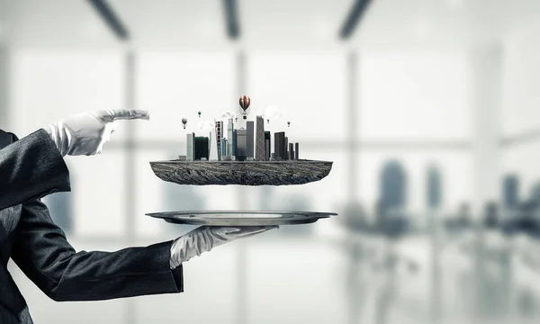 Cropped image of waitress's hand in white glove presenting modern city block on metal tray and pointing on it with office view on background. 3D rendering.