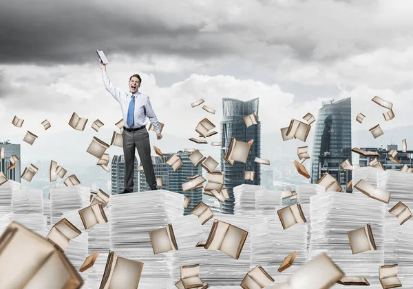 Businessman keeping hand with book up while standing among flying books with cityscape on background. Mixed media.