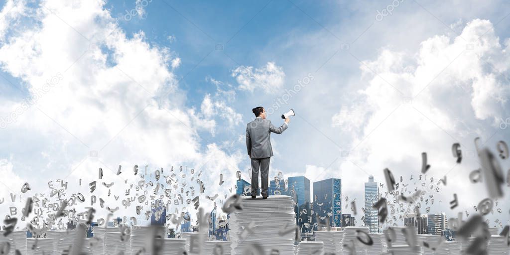 Businessman in suit standing among flying letters with speaker in hand and with skyscape on background. Mixed media.