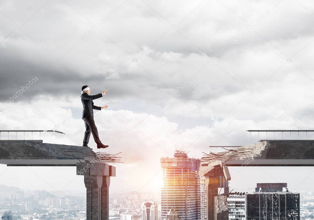 Businessman walking blindfolded on concrete bridge with huge gap as symbol of hidden threats and risks. Cityscape view with sunlight on background. 3D rendering.