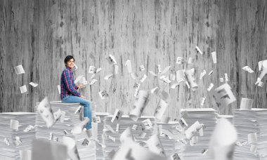 Kağıt belgeleri ile gri duvar arka plan üzerinde uçan arasında casual giyim oturan genç adam. Karışık teknik.