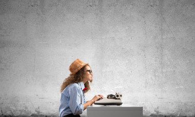 Genç güzel kadın yazar şapka ve gözlük arka planda gri beton duvara karşı masada otururken yazarak makinasıyla yan görünüm.