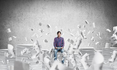 Kağıt belgeleri ile gri duvar arka plan üzerinde uçan arasında casual giyim oturan genç adam. Karışık teknik.