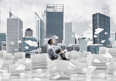 Düşünceli işadamı, arka planda şehir manzarası olan uçan kağıt uçaklar arasında bir yığın belgenin üzerinde otururken başka tarafa bakıyor. Karışık ortam.