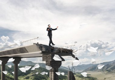 Gizli tehditler ve risklerin simgesi olarak beton köprüde uçan kağıt uçakların arasında gözü kapalı yürüyen bir işadamı. Arka planda gökyüzü ve doğa manzarası. 3B görüntüleme.