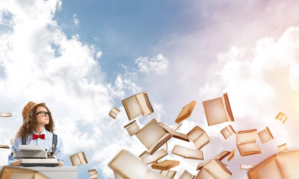 Young and beautiful woman writer in hat and eyeglasses using typing machine while sitting at the table among flying books with cloudy skyscape on background.