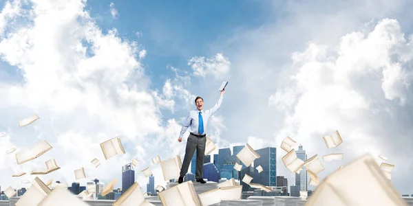 Businessman keeping hand with book up while standing among flying books with cloudly sky on background. Mixed media.